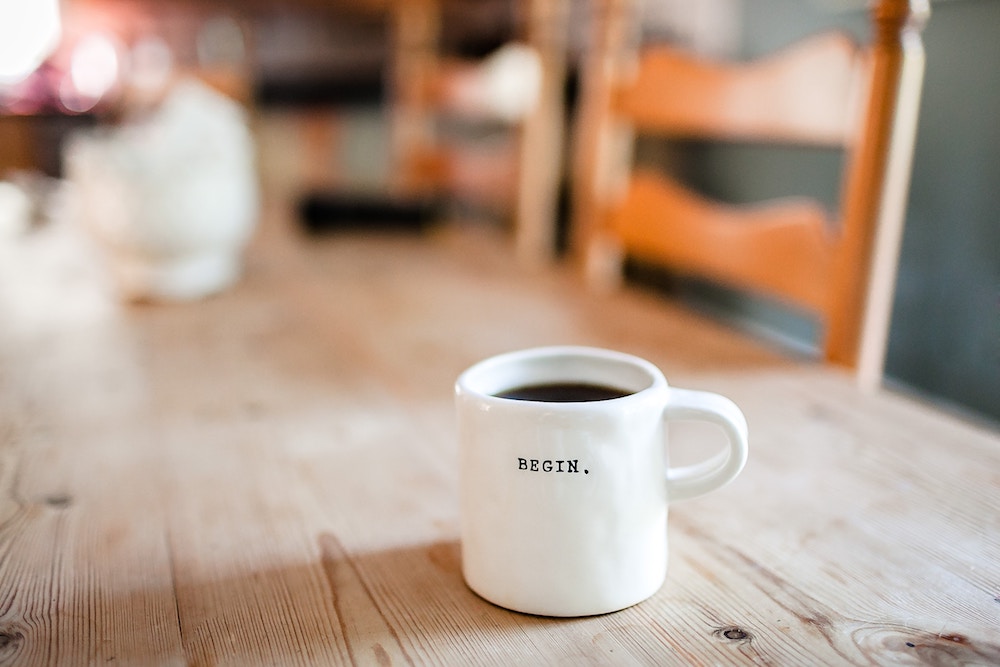 Coffee in white mug that says "begin" on a wooden table