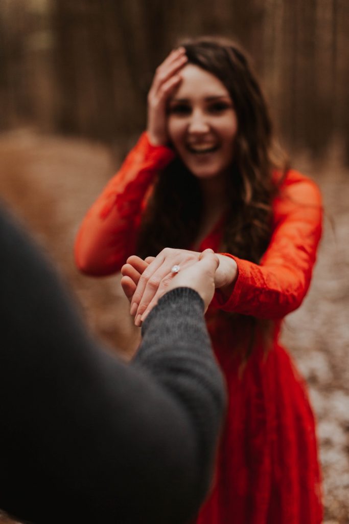 Girl in red dress holding hand of man with engagement ring on her finger, hand to head shocked