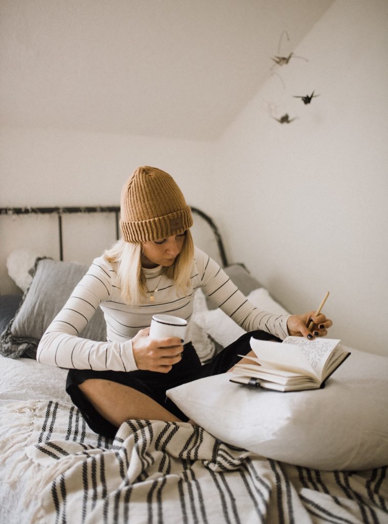 How to Interpret Your Dreams in 3 Steps | Happy As Annie | Best Life Book Club May selection is Steering by Starlight by Martha Beck, in which she explains how our intuition communicates with us through dreams. (Blonde woman in beanie holding coffee and jouranling in bed)