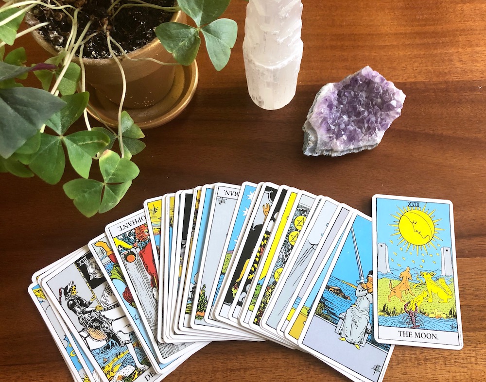 How to Break In and Cleanse Tarot Cards | Happy As Annie (The Moon and rest of Rider Waite tarot deck fanned across wooden table along with potted plant and crystals)