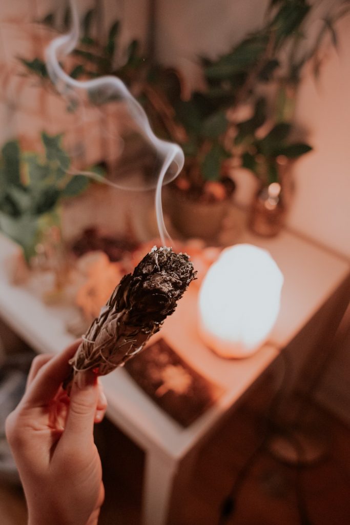 How to Cleanse a Space with Sage | Happy As Annie. How and why to burn sage to cleanse a space of negative energies. Practical tips and a step-by-step guide. #sage #burnsage #sagesmudge #sagewand (Smoking sage bundle in hand with table of potted plants and other objects blurred in background)