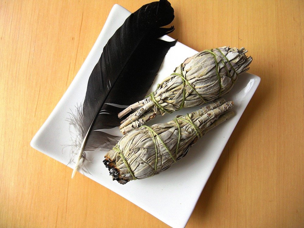 How to Cleanse a Space with Sage | Happy As Annie. How and why to burn sage to cleanse a space of negative energies. Practical tips and a step-by-step guide. #sage #burnsage #sagesmudge #sagewand (two sage bundles and black feather in white square dish on wooden table)