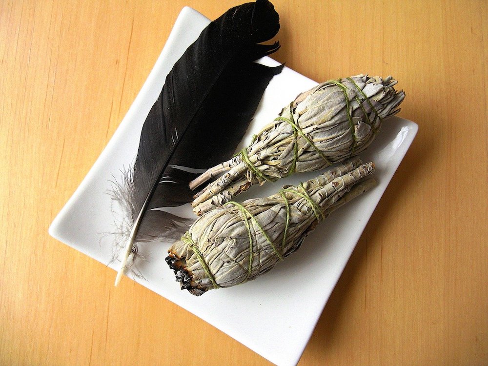 How to Cleanse a Space with Sage | Happy As Annie. How and why to burn sage to cleanse a space of negative energies. Practical tips and a step-by-step guide. #sage #burnsage #sagesmudge #sagewand (two sage bundles and black feather in white square dish on wooden table)