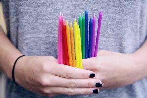 Girl in gray shirt and black nail polish holding several tall skinny rainbow colored candles in her hands