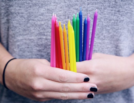 Girl in gray shirt and black nail polish holding several tall skinny rainbow colored candles in her hands