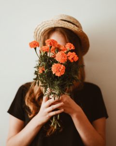 Woman wearing straw hat nad black tee holding orange flower bouquet in front of her face