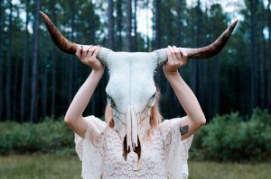 Girl in lace white dress holding up bull skeleton with horns in front of her face in woods