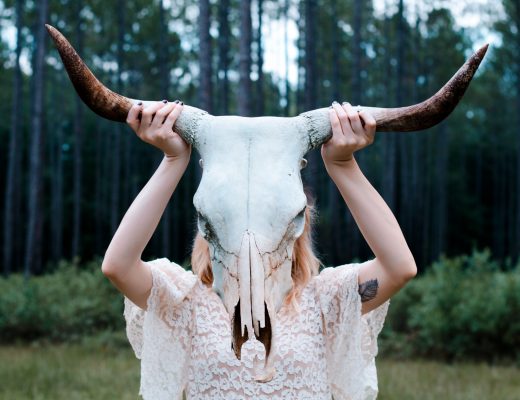 Girl in lace white dress holding up bull skeleton with horns in front of her face in woods