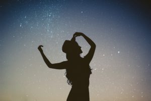 Woman's silhouette with arms up and wearing hat against starry sky background