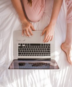Girl in pink pants on bed with white sheet typing on silver laptop
