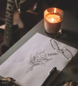 Candle in glass jar on table next to ink flower illustration and glasses