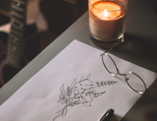 Candle in glass jar on table next to ink flower illustration and glasses